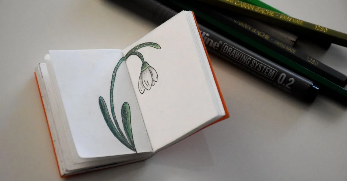 Mini carnet de croquis ouvert affichant un dessin de fleur de perce-neige avec des crayons de couleur et des stylos sur le côté.