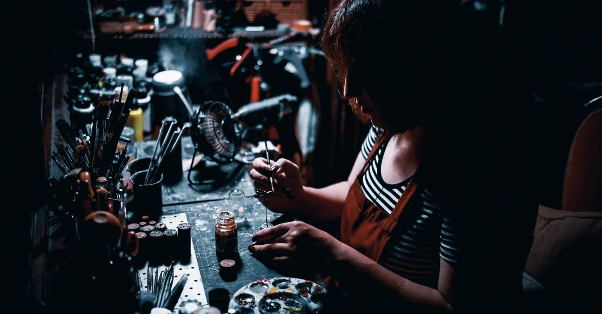Une femme artiste se concentre sur la peinture de modèles miniatures dans un atelier faiblement éclairé, rempli de pinceaux et de peinture.